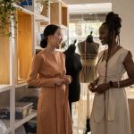 Dois designers de moda conversam sobre projetos em um estúdio com manequins e plantas ao fundo.