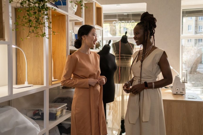 Dois designers de moda conversam sobre projetos em um estúdio com manequins e plantas ao fundo.